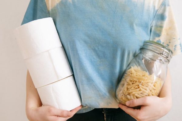 Woman in tie-dye shirt holding toilet paper rolls and pasta jar indoors.